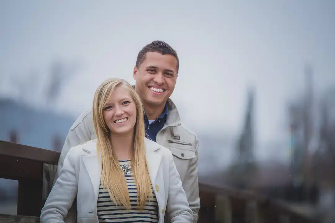 Two people stand close together outdoors, smiling at the camera on a cloudy day. The woman is in the foreground, and the man stands behind her. Both are wearing light-colored jackets.