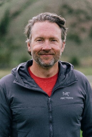 A man with short, greying hair and a beard smiles at the camera. He is wearing a dark blue Arc'teryx jacket over a red shirt, standing outdoors with a blurred natural backdrop.